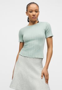 Νεαρή γυναίκα με πλεξούδες, φοράει εφαρμοστό ανοιχτοπράσινο τοπ με κοντά μανίκια και ανοιχτογκρι φούστα, στέκεται μπροστά σε ένα απλό φόντο.