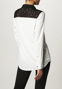 Blouse blanche avec empiècement en dentelle noire et col. Tissu doux, manches longues, poignets à un seul bouton, et ourlet arrondi. Motif en dentelle détaillé sur les épaules.