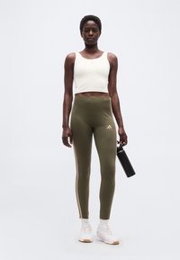 Αθλητική γυναίκα στέκεται με το βλέμμα προς τα εμπρός, φορώντας λευκό crop top, κολάν ελιάς με ρίγες, λευκά αθλητικά παπούτσια, κρατώντας μαύρο μπουκάλι νερού στο δεξί χέρι.