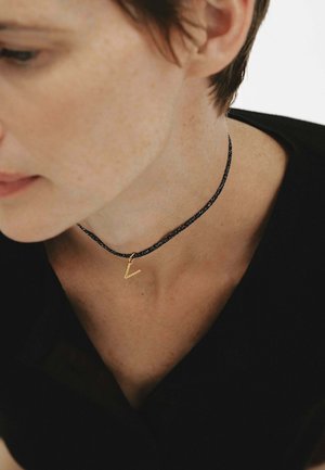 Primer plano de una persona que lleva un collar tipo choker de cuerda negra con un pequeño colgante dorado en forma de V, vestida con una camiseta negra.