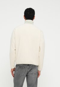 Lys beige fleecejakke med høj krave, ribstrikkede manchetter og en blød tekstur. Båret med grå denimjeans, der har baglommer.