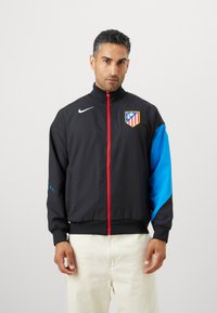 Mustmust ja sinine Nike jakk punase tõmblukuga, millel on meeskonna logo rinnal ja mustrilised aksessuaarid varrukatel.