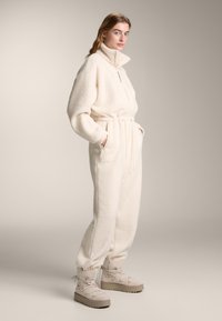 Kremno flis kombinezon z visokim ovratnikom, z zategovalnim trakom v pasu in elastičnimi manšetami. Kombiniran z bež platformnimi škornji. Gladek tekstura.