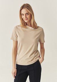 Beige īsām piedurknēm T-krekls ar mīkstu, gludu tekstūru un drapētu kakla izgriezumu, apvienots ar tumši ziliem biksēm. Vienkāršs, moderns dizains.
