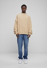 Beige oversized genser med høy hals, kombinert med lyseblå jeans og hvite sneakers. Genseren har senkede skuldre og en avslappet passform.
