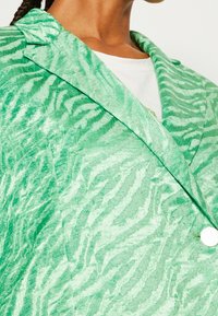 Blazer verde con textura y patrón de cebra, que presenta un cuello con muesca, un solo botón plateado y un tejido suave y aterciopelado.