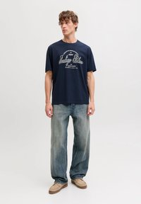 Μπλε ναυτικό t-shirt με λευκό κείμενο, σε συνδυασμό με ανοιχτό μπλε τζιν. Το μοντέλο στέκεται σε φωτεινό φόντο φορώντας καφέ μοκασίνια.