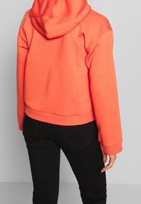 Krátká mikina s kapucí v jasně korálové barvě, vyrobená z měkké látky. Má prostornou kapuci a žebrované manžety, doplněná černými přiléhavými kalhotami.