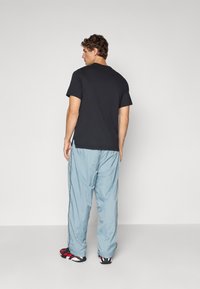 Mand, der står med ryggen til iført en sort t-shirt, lyseblå træningsbukser og sort, hvid og rød sneakers på en ensfarvet grå baggrund.