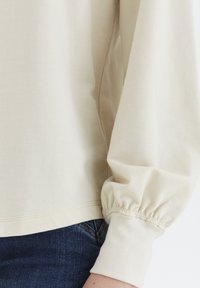 Beige hosszúujjú pulóver begyűrt mandzsettákkal és puha textúrával, sötétkék farmerral párosítva. Az egyszerű, minimalista dizájn jellemzi.