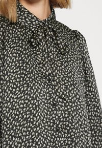 Blusa negra con patrón floral blanco, con cuello de lazo, cierre de botones y tejido texturizado, que destaca una silueta ajustada.