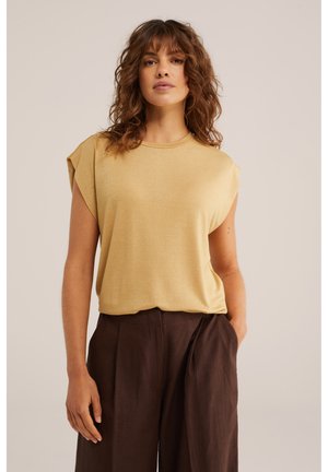 Femme aux cheveux bouclés portant un haut beige sans manches et un pantalon plissé taille haute marron foncé, debout avec une main dans la poche.