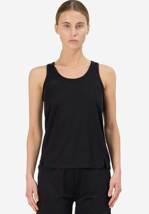 Femme portant un débardeur de sport noir sans manches et un pantalon noir, debout devant un fond clair uni.