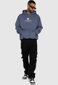 Sinine kapuutsiga särk, millel on esikott ja logo, koos mustade cargo-pükstega, millel on mitu taskut. Mudel kannab prille ja tenniseid.