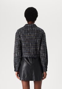 Giacca tweed tagliata con fili blu, neri e multicolori, dotata di colletto e maniche voluminose. Abbinata a una mini gonna in pelle nera.