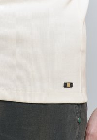 Lichtbeige geribde shirt met een zwart etiket met gele strepen aan de zoom; gecombineerd met donkergrijze stoffen broek en een knoopdetail.