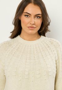 Maglione crema lavorato a maglia con lavorazione a costine testurizzate e accenti di perle sul giromanica, con scollo rotondo e maniche lunghe.