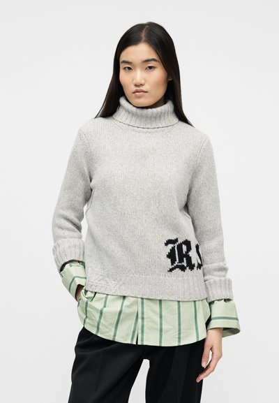 Γυναίκα με μακριά σκούρα μαλλιά που φοράει ανοιχτόγρι ζιβάγκο πάνω από πράσινο ριγέ πουκάμισο και μαύρο παντελόνι, στέκεται μπροστά σε ένα απλό φόντο.