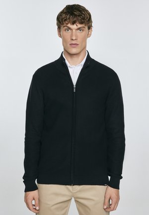Zwarte zip-up trui met een textuur gebreid ontwerp, hoge kraag en geribde manchetten, gedragen over een wit shirt. Strakke lijnen en een getailleerde silhouet.