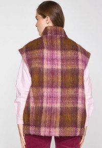Gilet en laine avec un large col, motif tartan violet, rose et marron, d'une texture douce, porté sur une chemise rayée.