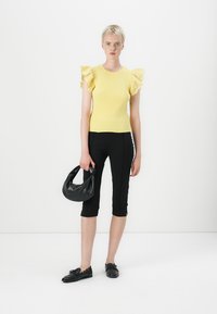 Gul ribbet strikket topp med volangerm, kombinert med svarte avkuttede leggings. Modellen holder en svart teksturert håndveske og har på seg svarte slip-on sko.