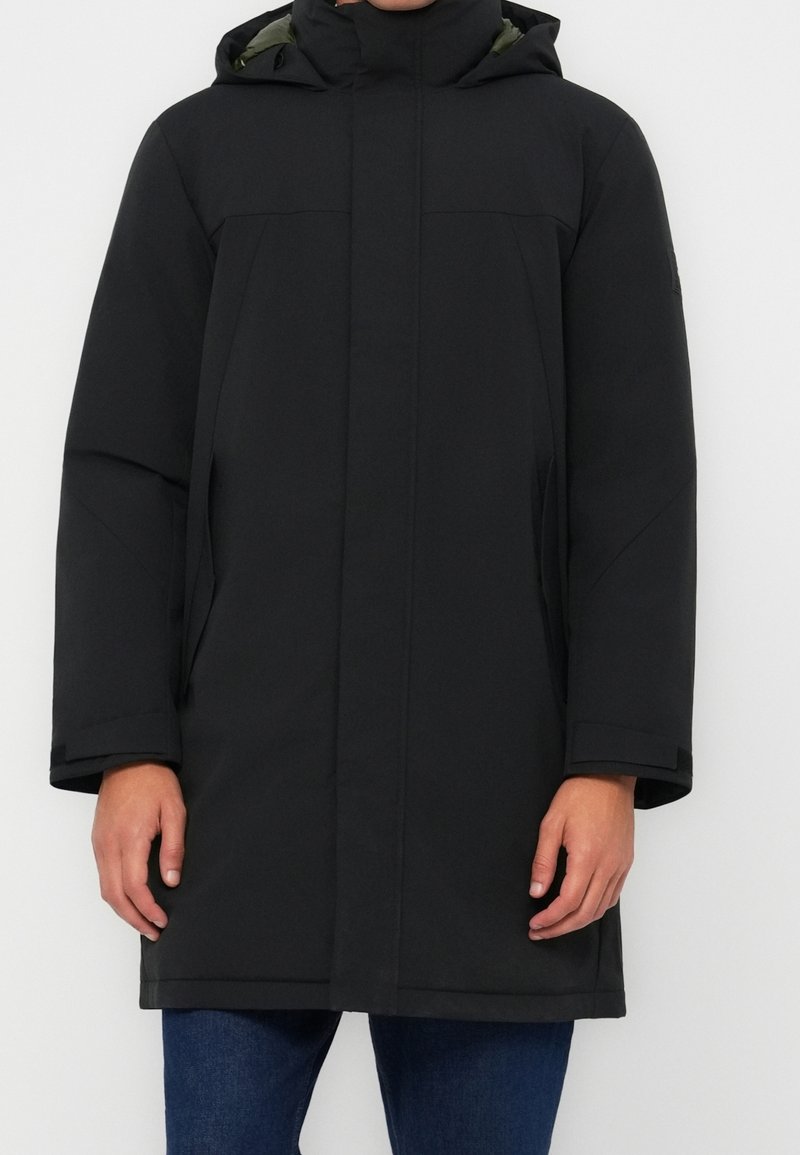 Homme portant un manteau d'hiver noir à capuche avec fermeture éclair frontale dissimulée et poches latérales, associé à un jean bleu.
