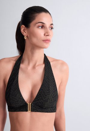 Femme aux cheveux foncés attachés, portant un haut de bikini noir à licou avec un fermoir doré et des boucles d'oreilles géométriques dorées, regardant vers la droite.