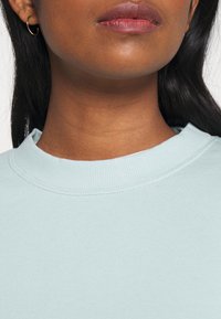 Ljusblå sweatshirt med ribbad rund halsringning, som har en mjuk textur och minimalistisk design. Visar en närbild av krageområdet.