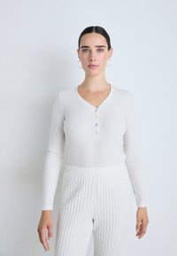 Vit långärmad henley-topp med V-ringning, försedd med fyra knappar framtill. Kombinerad med ljusa, texturerade rivetter, vilket ger en enhetlig ljus look.