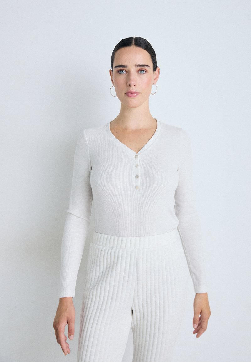 Vit långärmad henley-topp med V-ringning, försedd med fyra knappar framtill. Kombinerad med ljusa, texturerade rivetter, vilket ger en enhetlig ljus look.