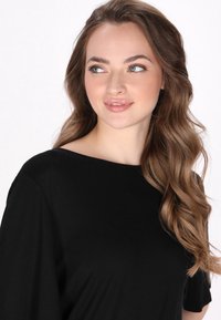 Sort kortærmet top med bådudskæring og blød, glat tekstur. Modellen har lyst bølget hår og et neutralt makeup-look.