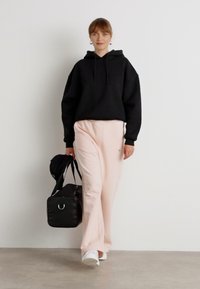 Haut à capuche noir surdimensionné, pantalon de survêtement rose clair, baskets blanches et sac de sport noir. Textures lisses avec un branding subtil sur le pantalon.