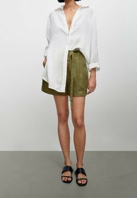 Camicia bianca di lino con bottoni e maniche lunghe; abbinata a pantaloni corti di lino verdi con cintura a fiocco. Sandali neri da infilare.