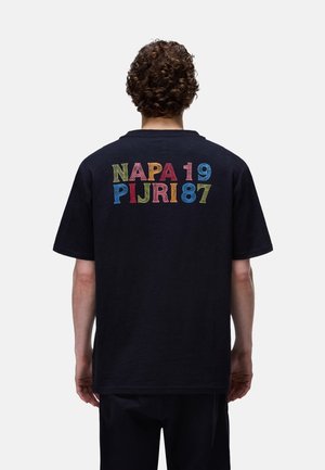 Person mit lockigem Haar trägt ein schwarzes T-Shirt mit buntem Text "NAPA 19 PIJRI 87" auf dem Rücken vor einem schlichten weißen Hintergrund.