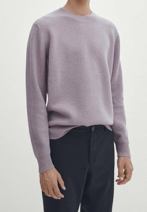 Man wearing a light purple textured crewneck sweater and dark trousers against a plain white background.