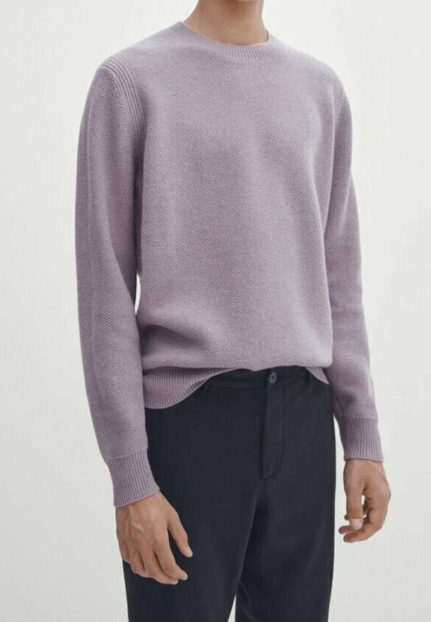 Homme portant un pull texturé violet clair à col rond et un pantalon foncé sur un fond blanc uni.