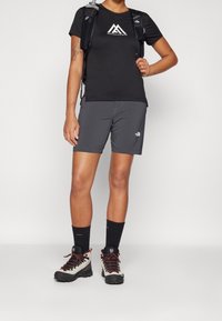 Musta lyhythihainen urheilupaita, jossa valkoinen logo, yhdistetty harmaisiin shortseihin. Käytetty vaelluskengillä ja tummilla sukilla, mukana reppu.