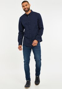 Marineblaue Hemdbluse mit kleinen weißen Punkten, langen Ärmeln und Kragen, kombiniert mit dunkelblauen Jeans und schwarzen Schuhen.