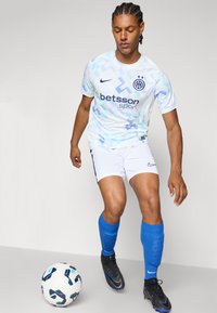 Maillot de football blanc avec des motifs géométriques bleu clair et un logo, associé à un short blanc et des chaussettes bleu marine jusqu'aux genoux. Chaussures de foot noires. Un ballon de football à proximité.