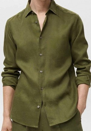 Homme portant une chemise olive à manches longues boutonnée avec col et manches retroussées, les mains partiellement dans les poches, sur fond uni.