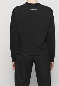 Felpa nera in tessuto morbido con scollo rotondo, maniche lunghe e polsini a costine. Presenta un piccolo logo bianco sulla parte superiore della schiena.