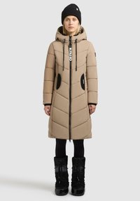 Beige pufferjacka med huva, med diagonalt quiltat mönster, dragkedja fram, svarta detaljer på fickorna och en logotags. Bärs med svarta stövlar.