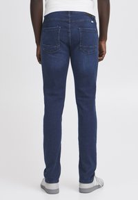 Donkerblauwe denim jeans met een rechte pasvorm, voorzien van twee achterzakken en subtiele stikdetails, gecombineerd met grijze sneakers.