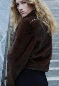 Brązowa kurtka z futra syntetycznego o miękkiej fakturze, z wysokim kołnierzem i luźnym krojem. Modelka pozuje na tle kamiennej ściany.