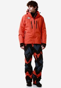 Giacca impermeabile arancione con zip nere e logo, abbinata a pantaloni neri con un motivo geometrico rosso e grigio.