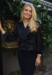 Czarna satynowa bluzka z bufiastymi rękawami i wzorem w kropki, zestawiona z dopasowaną czarną spódnicą. Srebrna biżuteria i luksusowy detal tła.