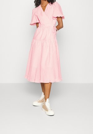 Vrouw draagt een midi wikkeljurk met roze en witte strepen, gerimpelde mouwen en zoom, gecombineerd met beige hoge sneakers.