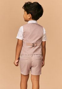 Gilet beige clair avec un détail de boucle dans le dos, associé à une chemise blanche à motif floral et un short beige clair assorti.