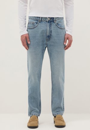 Hombre vestido con vaqueros de pierna recta azul claro, mocasines beige y una camisa blanca de manga larga, de pie contra un fondo liso.