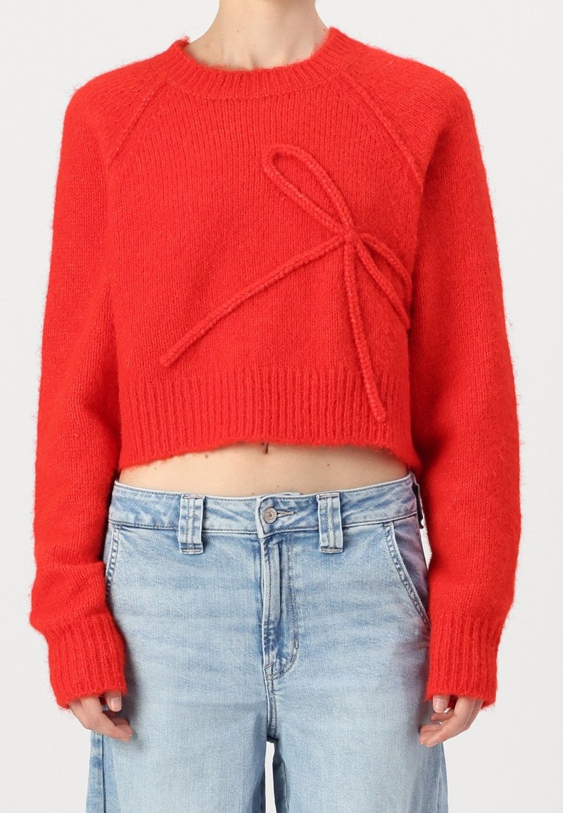 Suéter corto rojo hecho de suave tejido de punto, con un destacado detalle de lazo en la parte delantera y puños y dobladillo acanalados. Combinado con jeans claros.